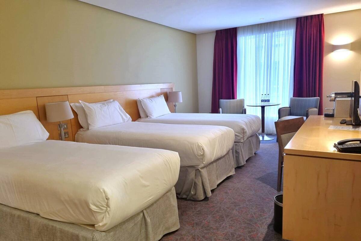 A hotel room with two beds, a desk, a chair, a table, and a radiator.