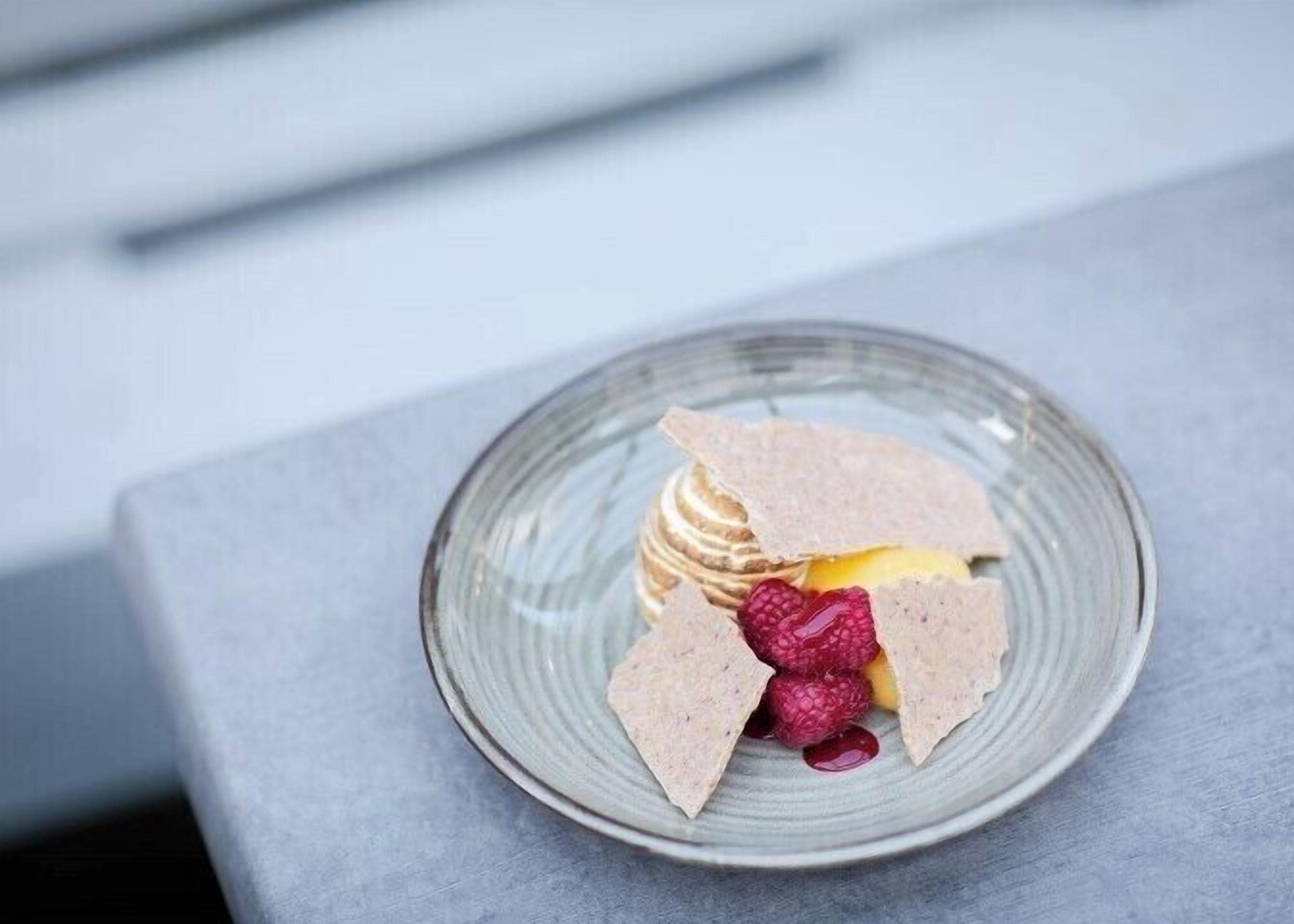 Un plato de postre con una porción de pastel, frambuesas y una fruta sobre una superficie rugosa.