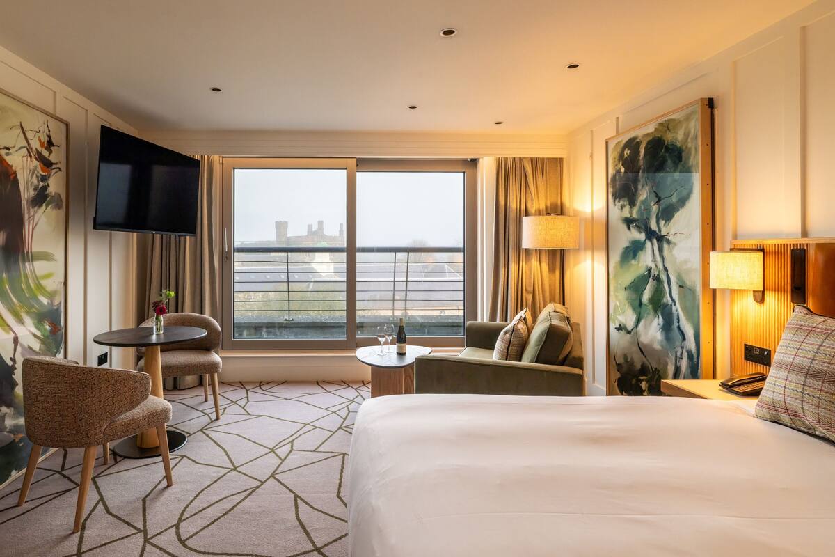 A hotel room with a bed, a sofa, a chair, a small table with a bottle and glass, and a TV.