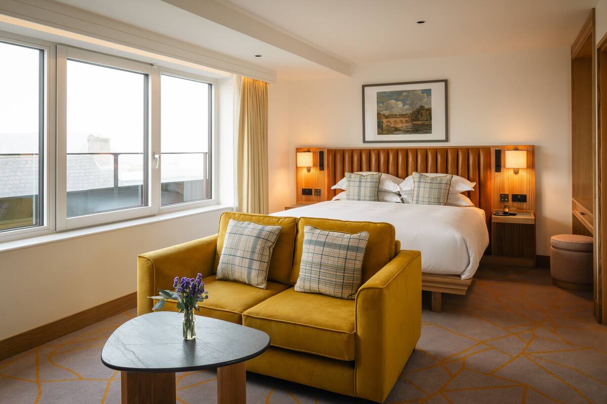 A hotel room with a bed, a sofa, a table with flowers, a window with curtains, and a painting on the wall.
