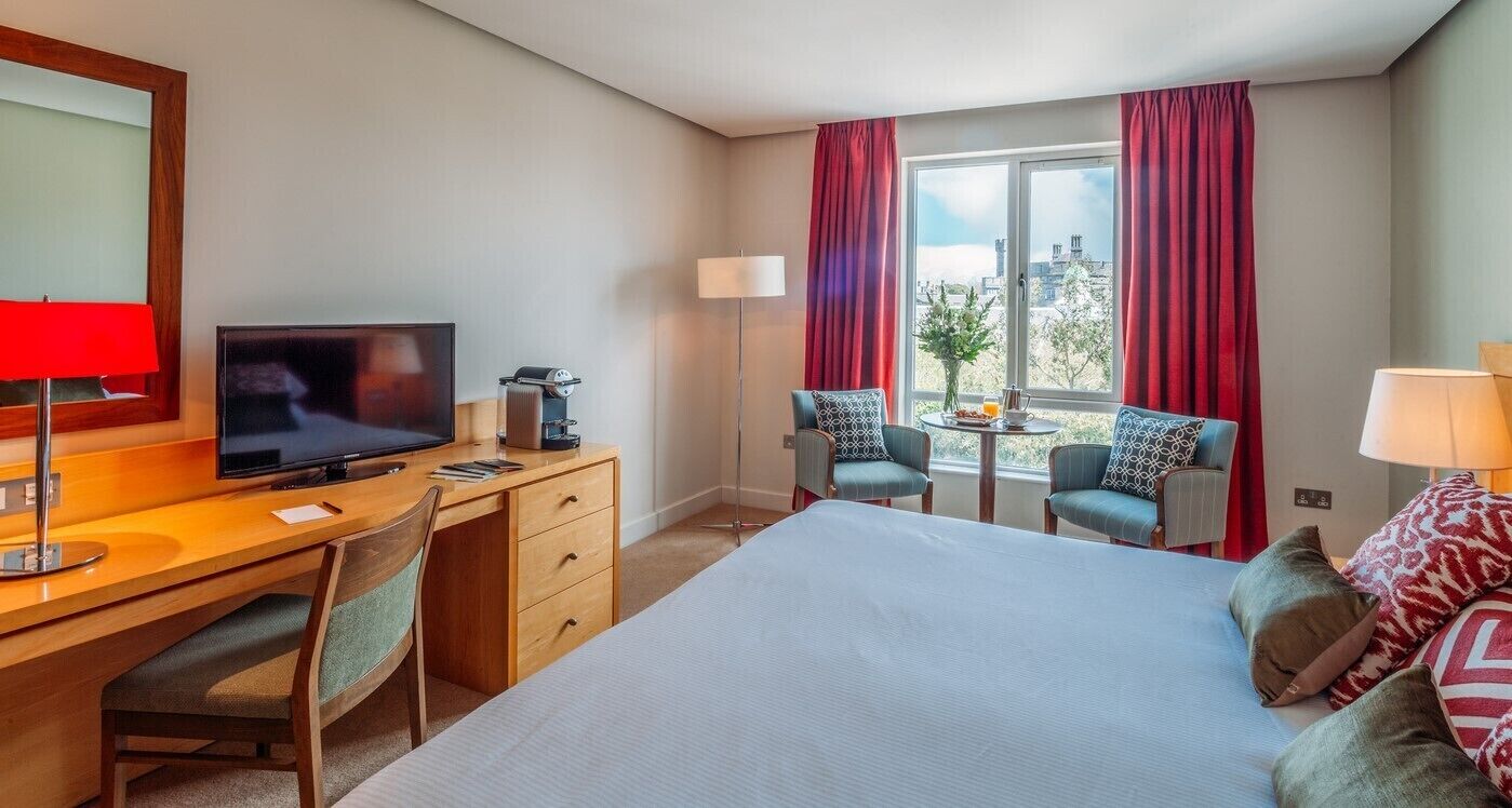 Habitación de hotel con cama, escritorio con televisor, cafetera, dos sillas, una mesita y ventana con cortinas.