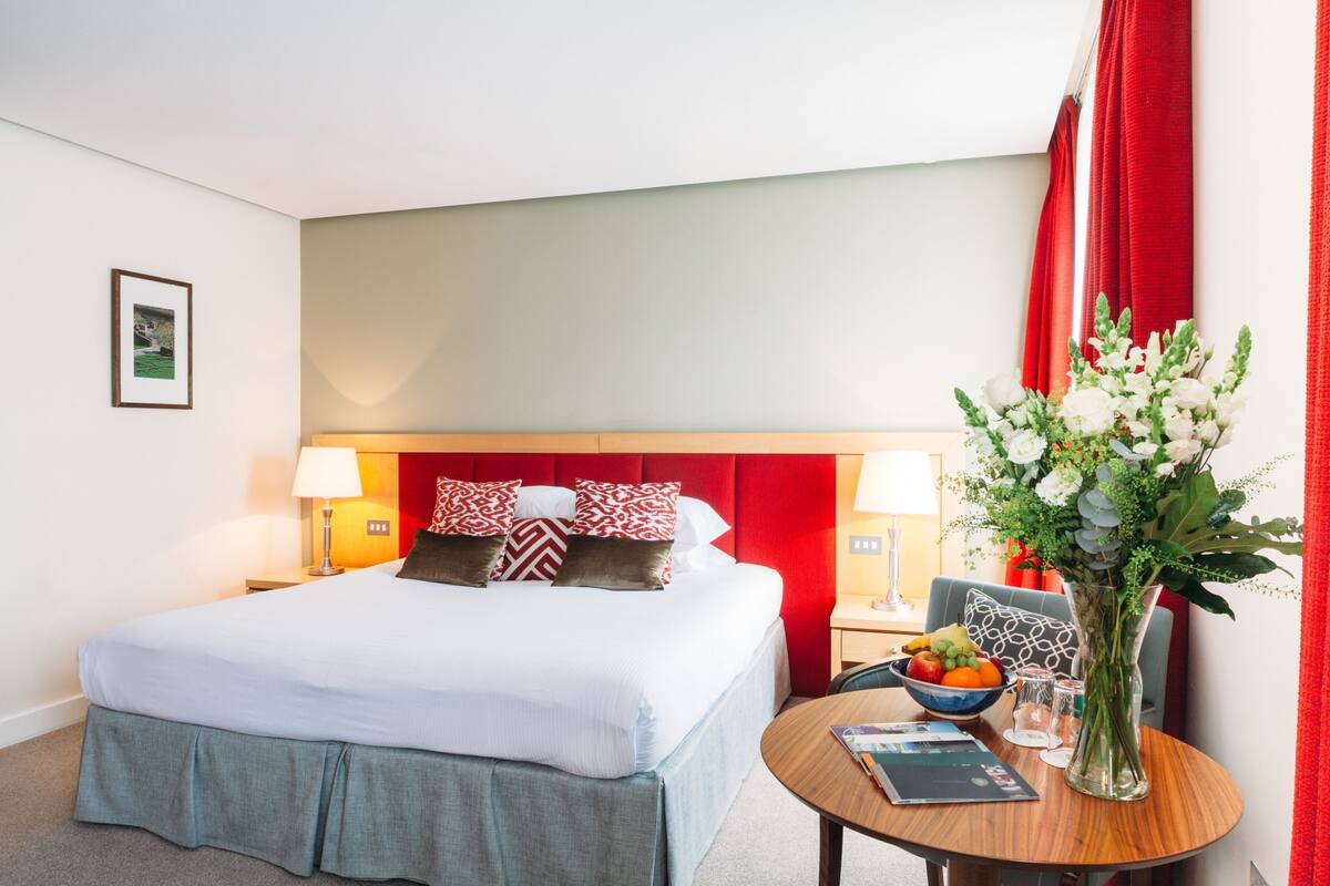 A hotel room with a bed, bedside tables, a round table with a vase of flowers, and a framed picture on the wall.