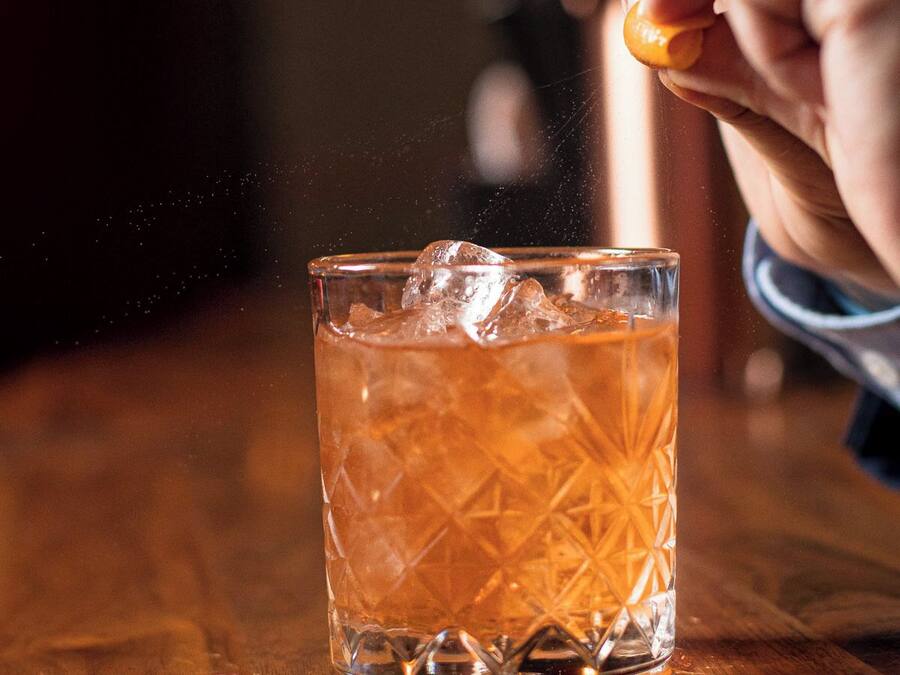 A hand squeezing an orange peel over a glass of amber-colored drink with ice cubes.