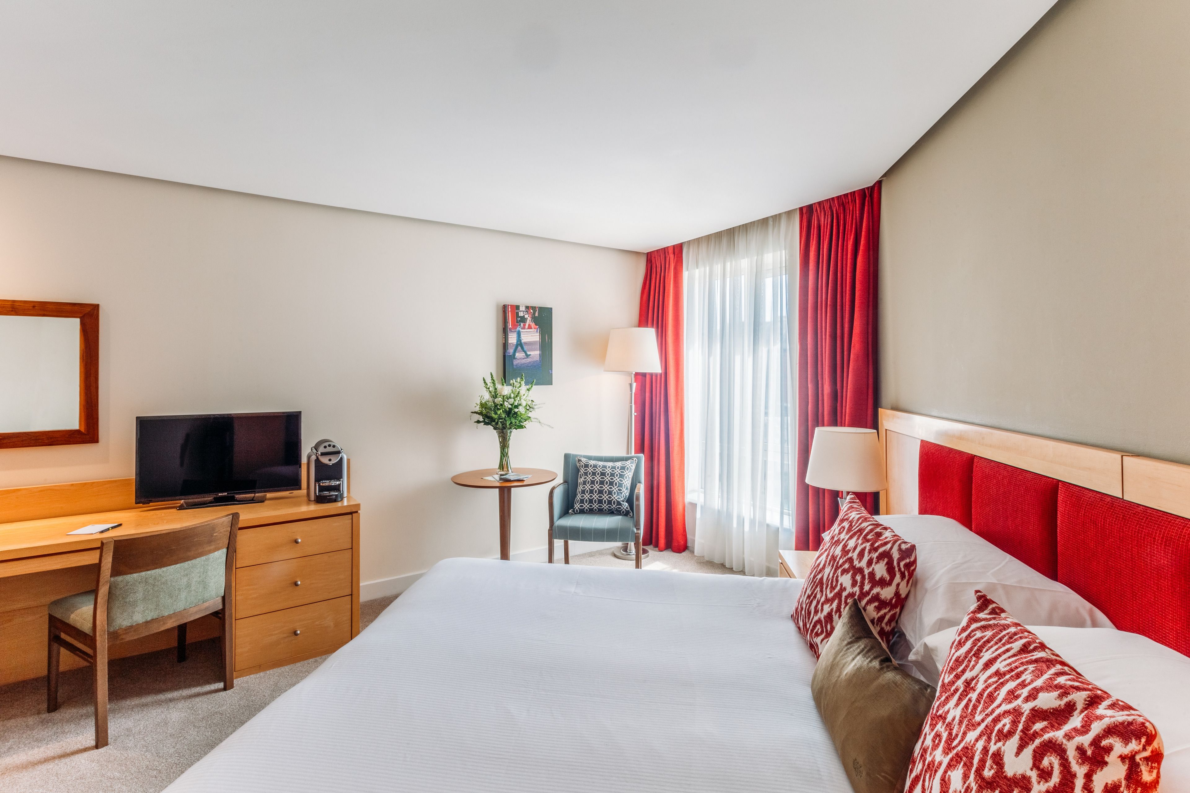 Una camera d'albergo con un letto, una scrivania con televisore, una sedia, un tavolino con lampada e una finestra con tende rosse.