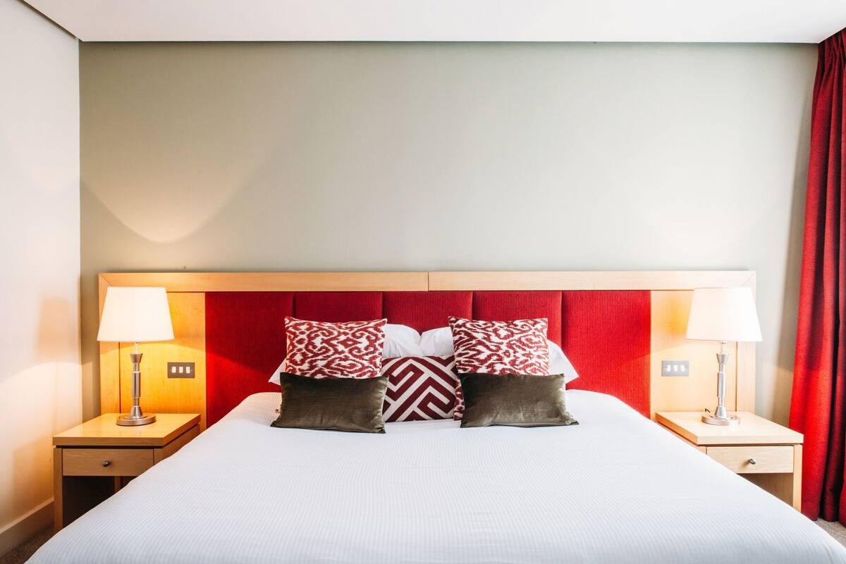 A hotel room with a large bed, two bedside tables with lamps, and a red headboard.