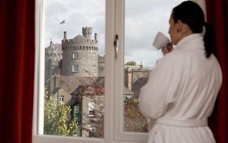 Una persona vestida con bata de baño frente a una ventana con vistas a un castillo.