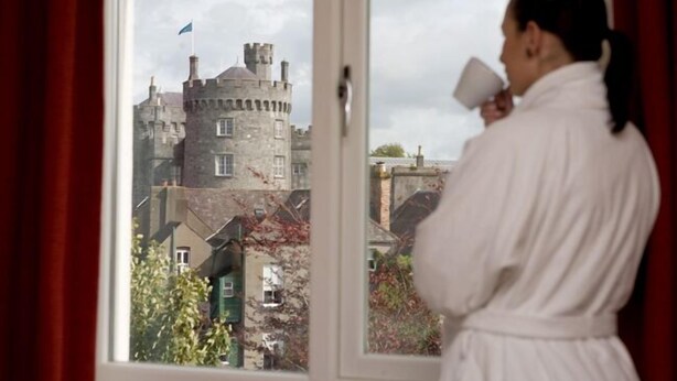 Eine Person in einem Bademantel steht an einem Fenster mit Blick auf ein Schloss.