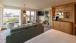 Ein Hotelzimmer mit einem großen Bett, einer Sitzecke mit Sofas und Sesseln, einem kleinen runden Tisch, einem an der Wand montierten Fernseher und einer Minibar mit Flaschen und Gläsern.