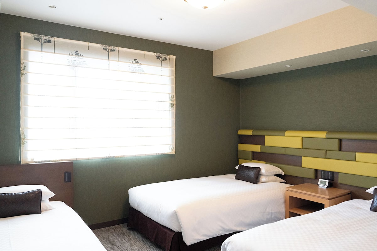 A hotel room with two beds, a window with blinds, a bedside table with a clock, and a headboard with a geometric design.