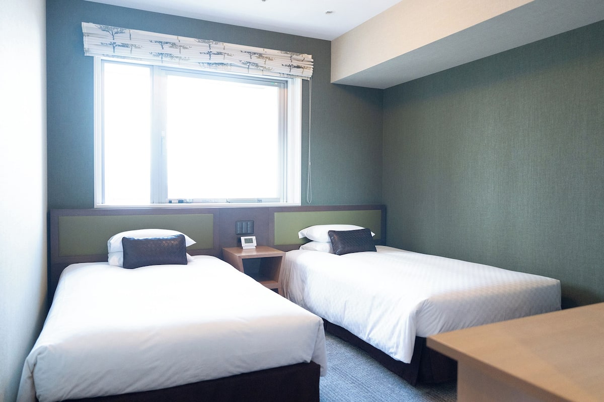 A room with two single beds, a wooden nightstand with a lamp, and a window with a curtain.
