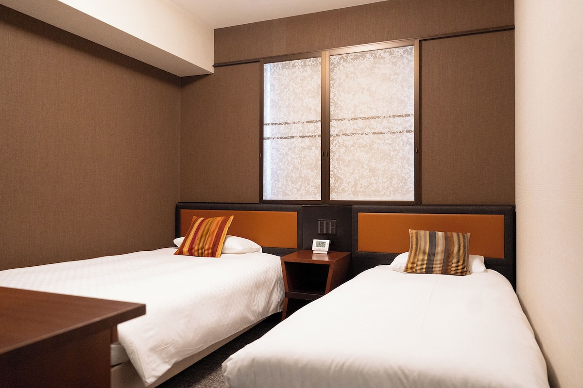 A hotel room with two single beds, a nightstand with a clock, and a window with a view.