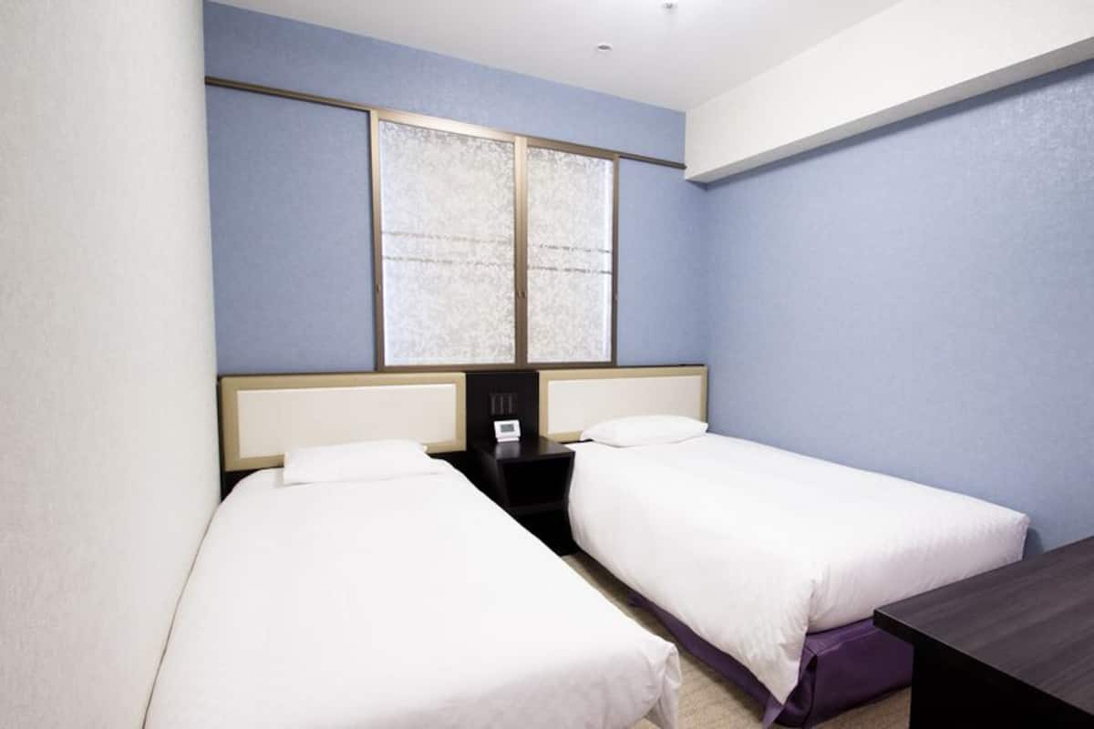 A hotel room with two beds, a small table, a nightstand, and a window.