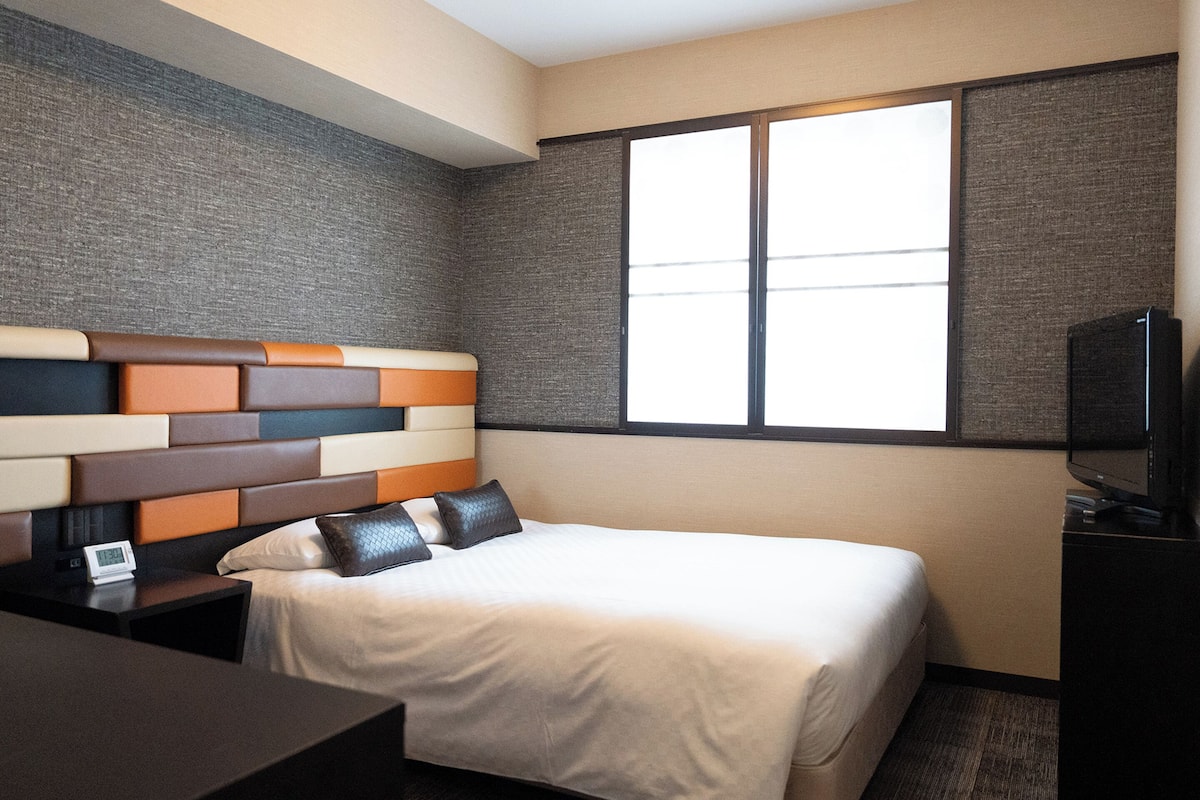 A hotel room with a modern bed, a flat-screen TV, and a large window providing natural light.