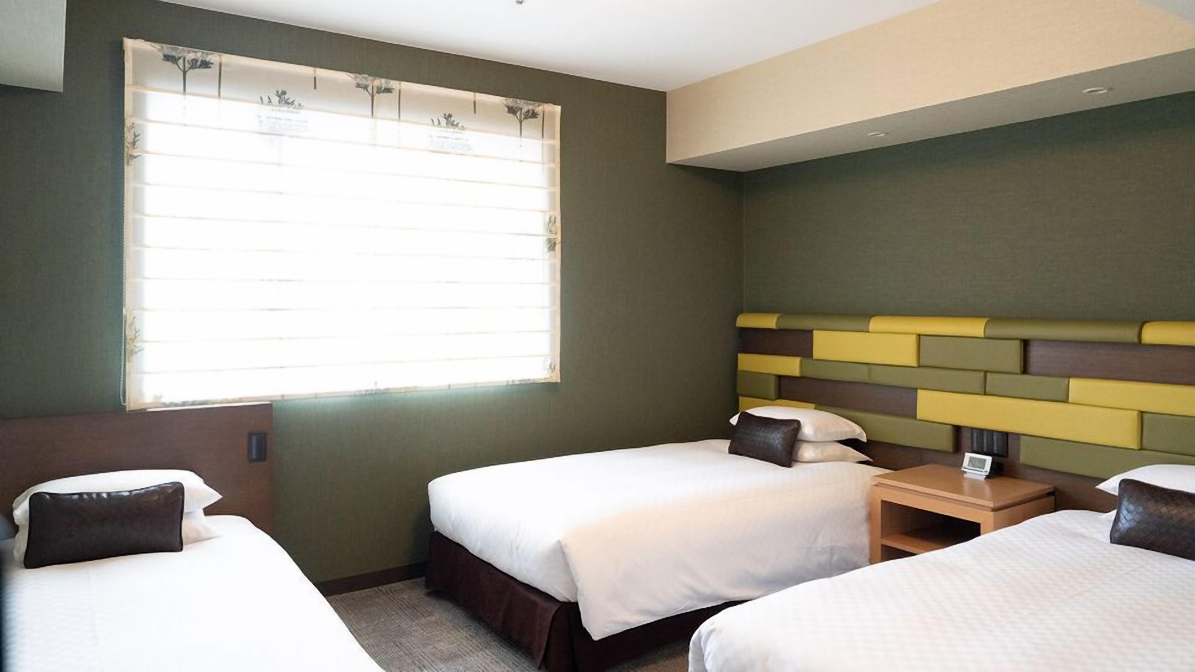 Une chambre d’hôtel avec deux lits, une fenêtre avec des stores, une table de chevet avec une lampe et une tête de lit ornée d’un motif géométrique.