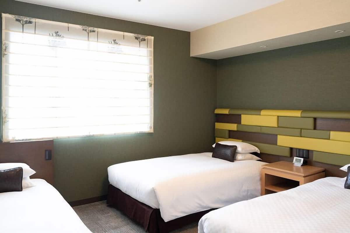 A hotel room with two beds, a window with blinds, a bedside table with a lamp, and a headboard with a geometric design.