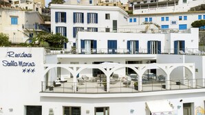 Un bâtiment blanc avec des volets bleus, une enseigne indiquant « Résidence Stella Marina », et des espaces extérieurs aménagés avec des tables et des chaises.