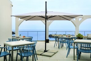 An outdoor patio with tables and chairs, a large umbrella, and a view of the sea. - Stella Marina (Ustica)