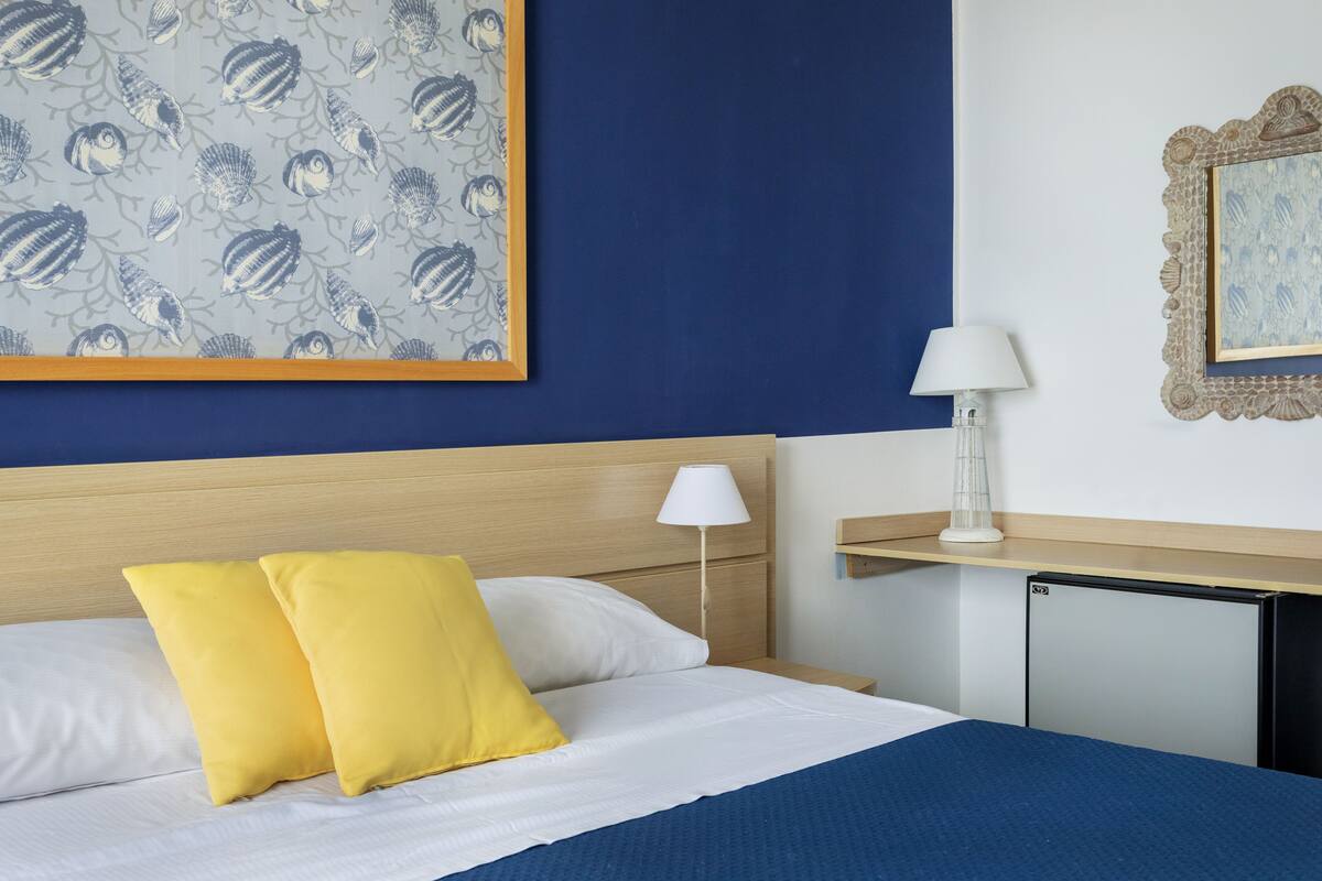 A neatly made bed with two pillows, a bedside lamp, a small desk with a lamp, and a framed picture on the wall.