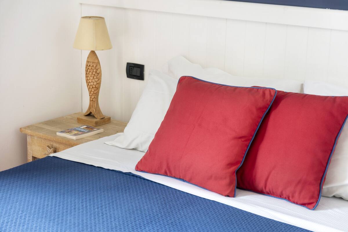 A neatly made bed with a blue bedspread, two red pillows with blue trim, a white pillow, and a wooden nightstand with a lamp and a book.