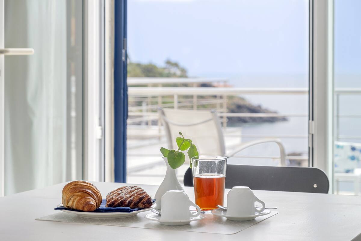 A table with croissants, a glass of tea, and a white vase with a green plant.