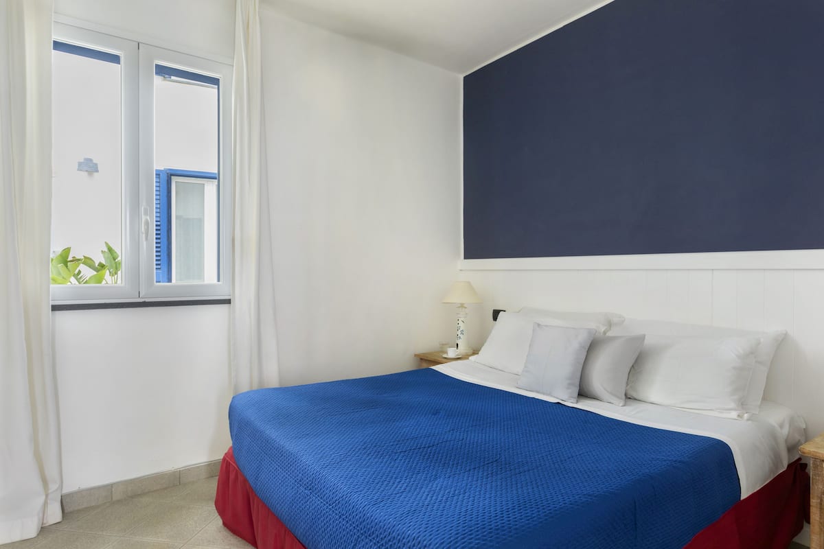 A bedroom with a bed, white curtains, a window, a nightstand, and a lamp.