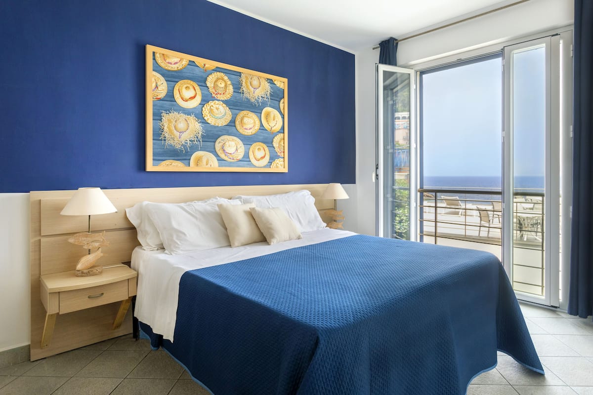A hotel room with a large bed, a nightstand, a lamp, and a view of the sea through a glass door.