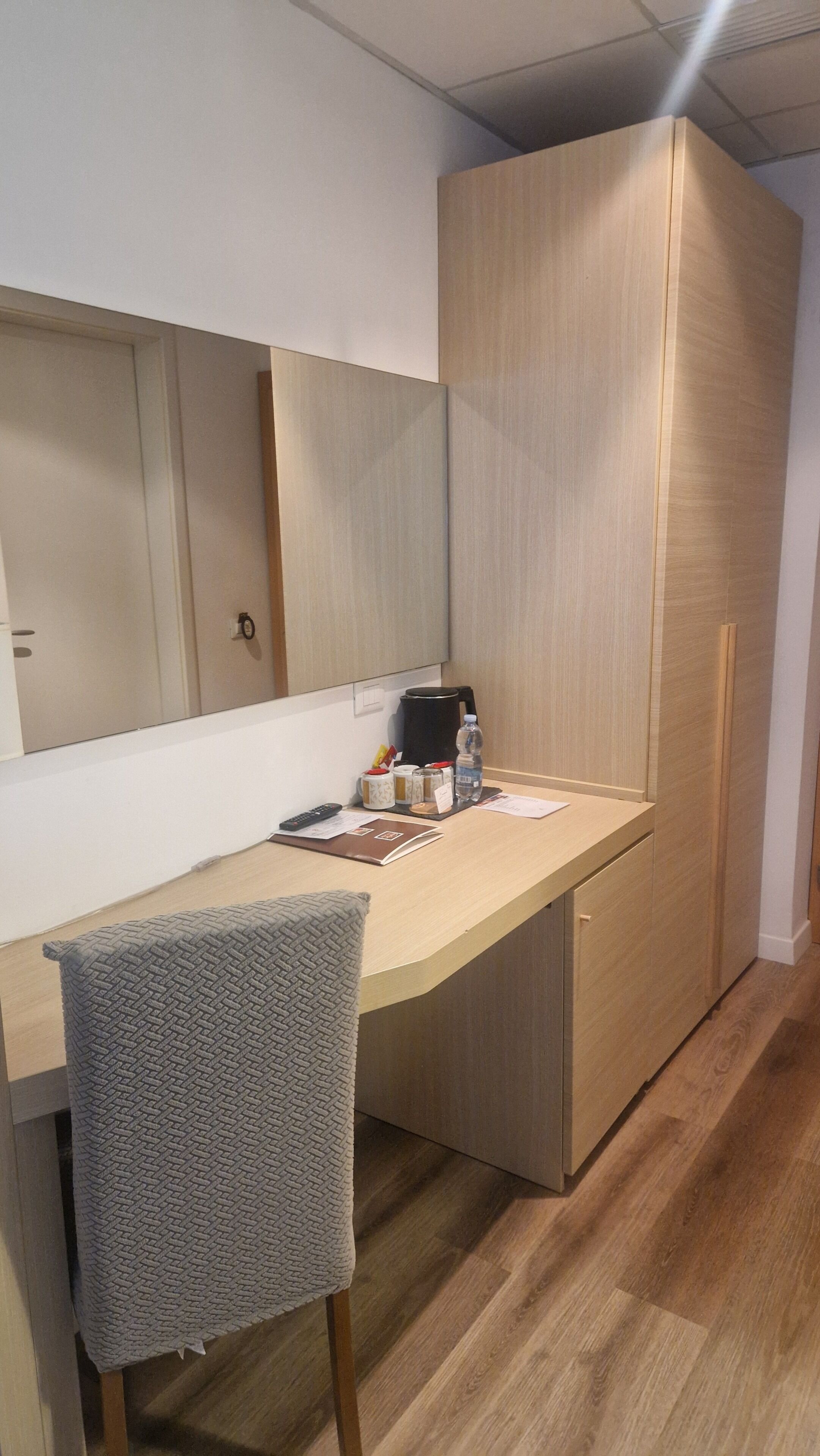 Camera d'albergo compatta con una scrivania in legno, una sedia, uno specchio, una macchina del caffè e una bottiglia d'acqua.