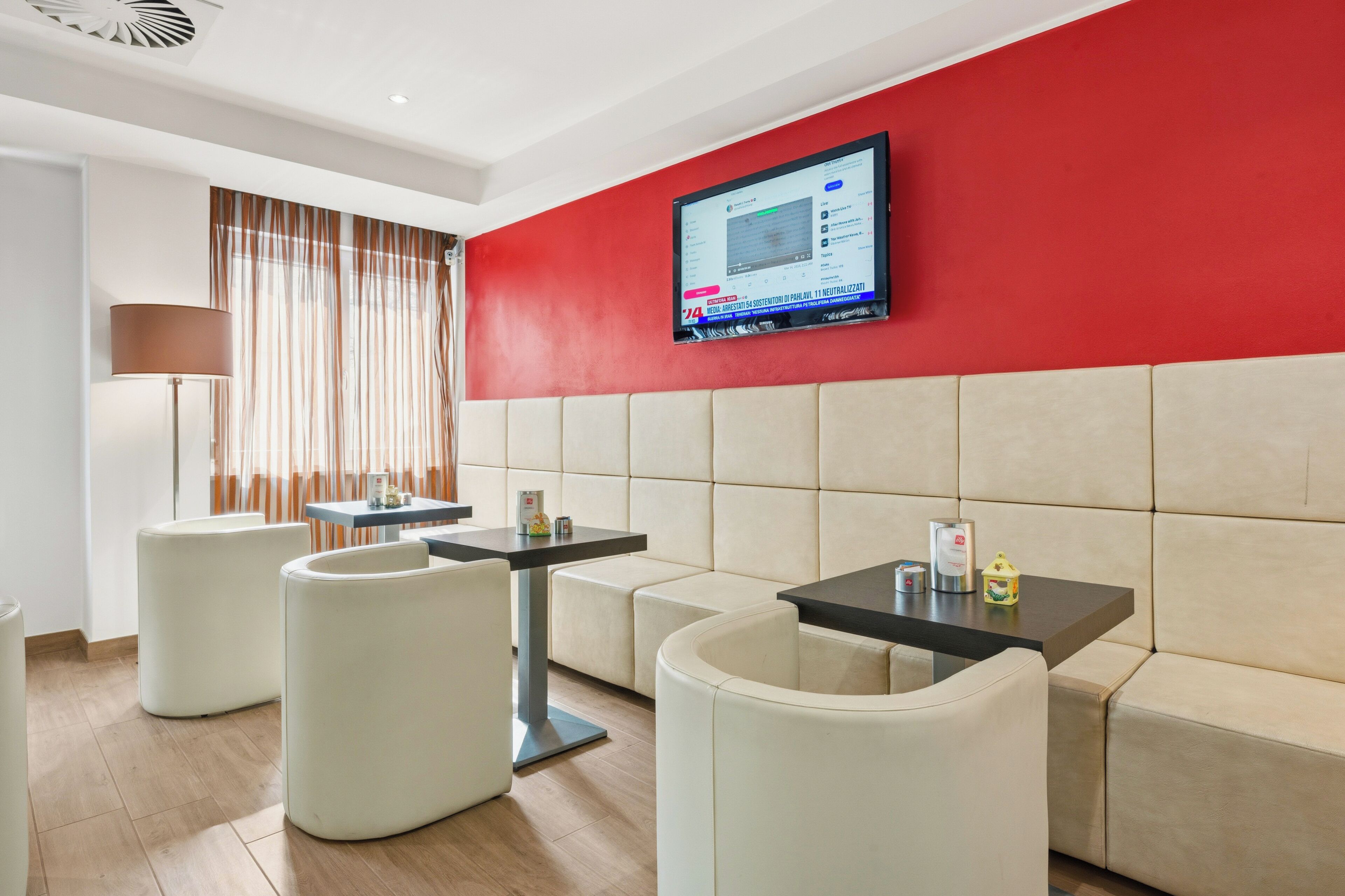 Ein modernes Hotelzimmer mit roter Akzentwand, einem flachbildfernseher über einer beigefarbenen gepolsterten Bank und einem kleinen Tisch mit Lampe.