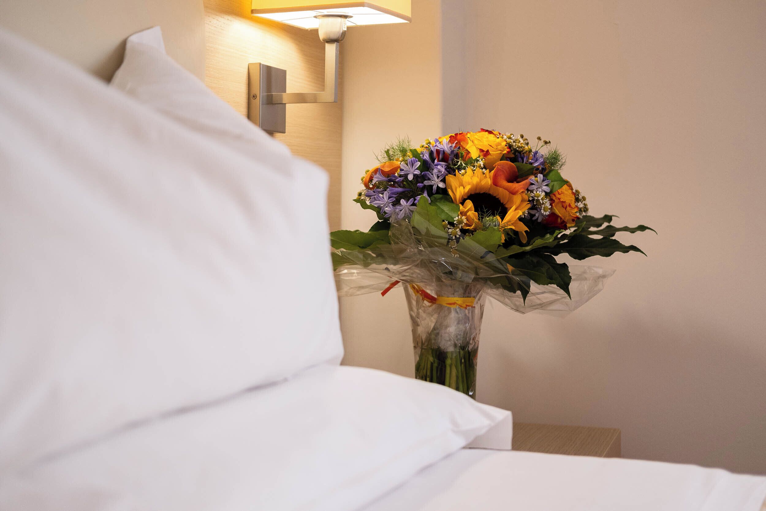 A hotel room with a bed, white bedding, a nightstand with a vase of flowers, and a wall-mounted lamp.