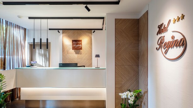Un mostrador de recepción de hotel con diseño moderno, un letrero que dice 'Hotel Regina' y una zona de recepción con un banco y plantas en macetas.