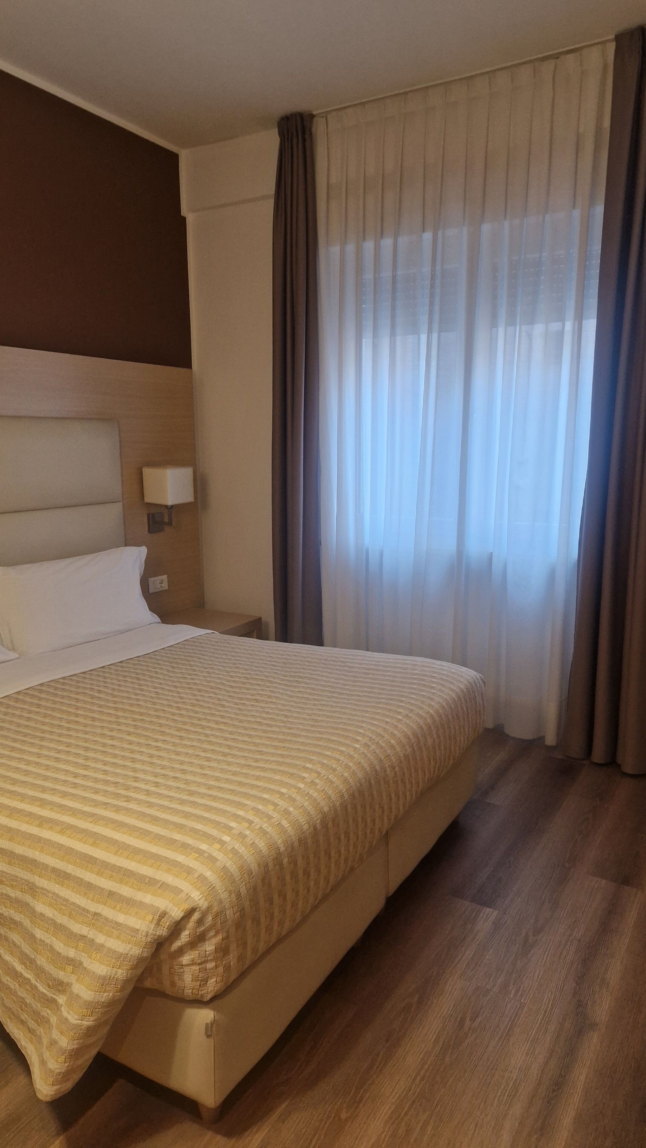 Una camera d'albergo con un letto, un comodino, una lampada e una finestra con tende.