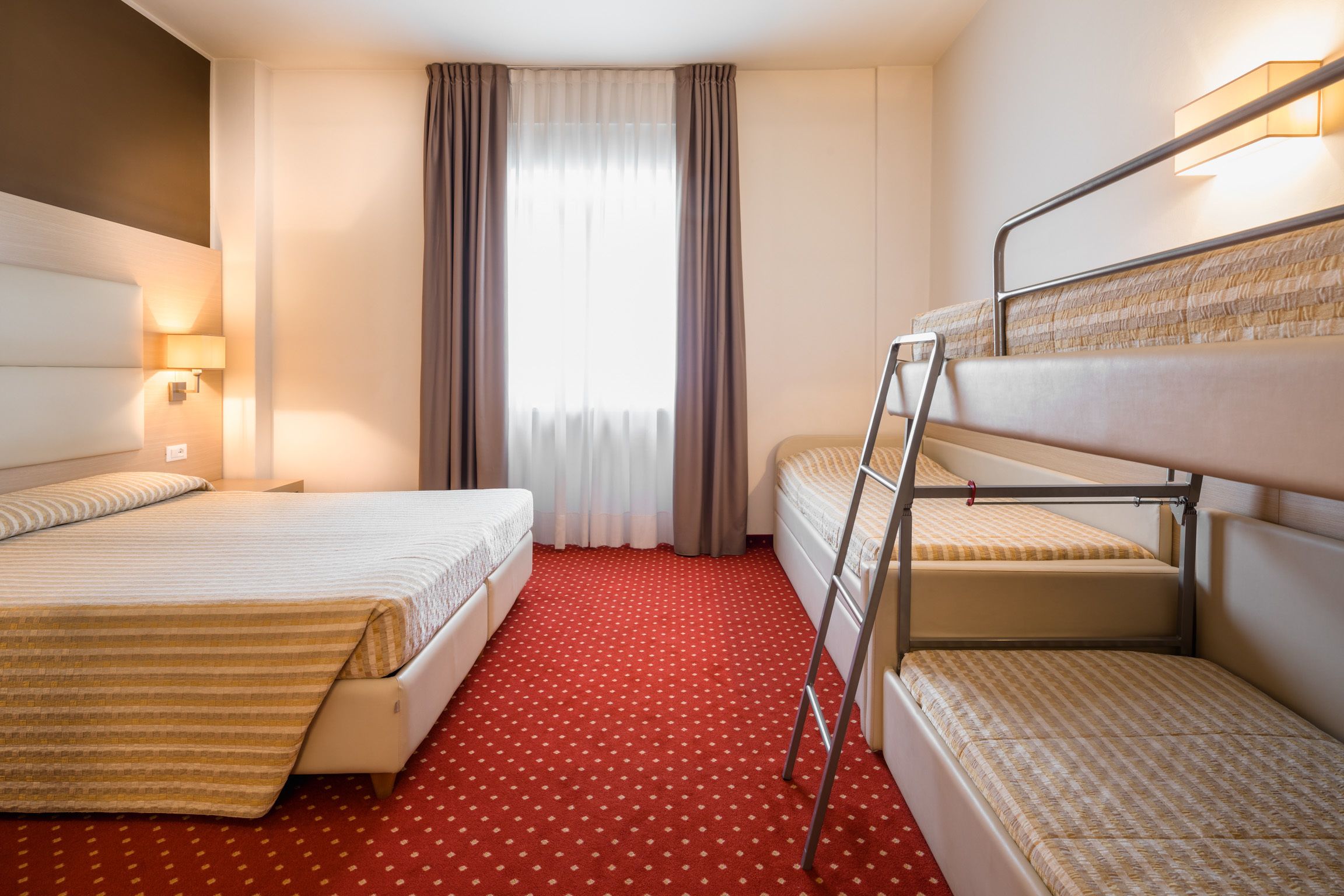 Habitación de hotel con dos camas, alfombra roja, ventana con cortinas y una lámpara fijada a la pared.
