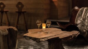 Een rustieke houten tafel met twee glazen witte wijn en een kaars, voor een stenen muur, met houten stoelen op de achtergrond.