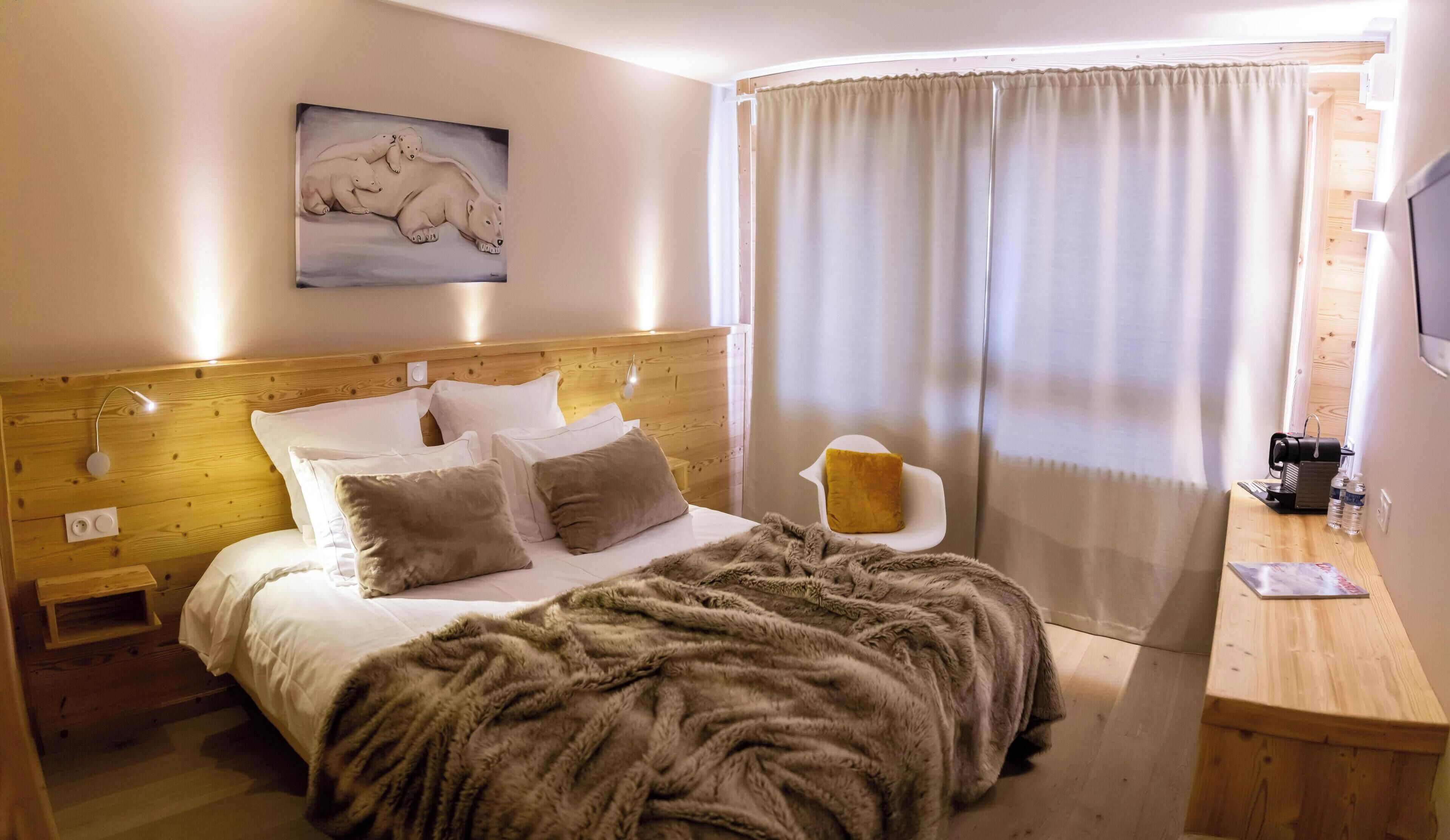 Habitación de hotel con cabecera de madera, cama con ropa de cama blanca y marrón, manta de piel, pintura de osos polares, mesita de noche de madera, silla con cojín amarillo, escritorio de madera con cafetera y ventana con cortinas.