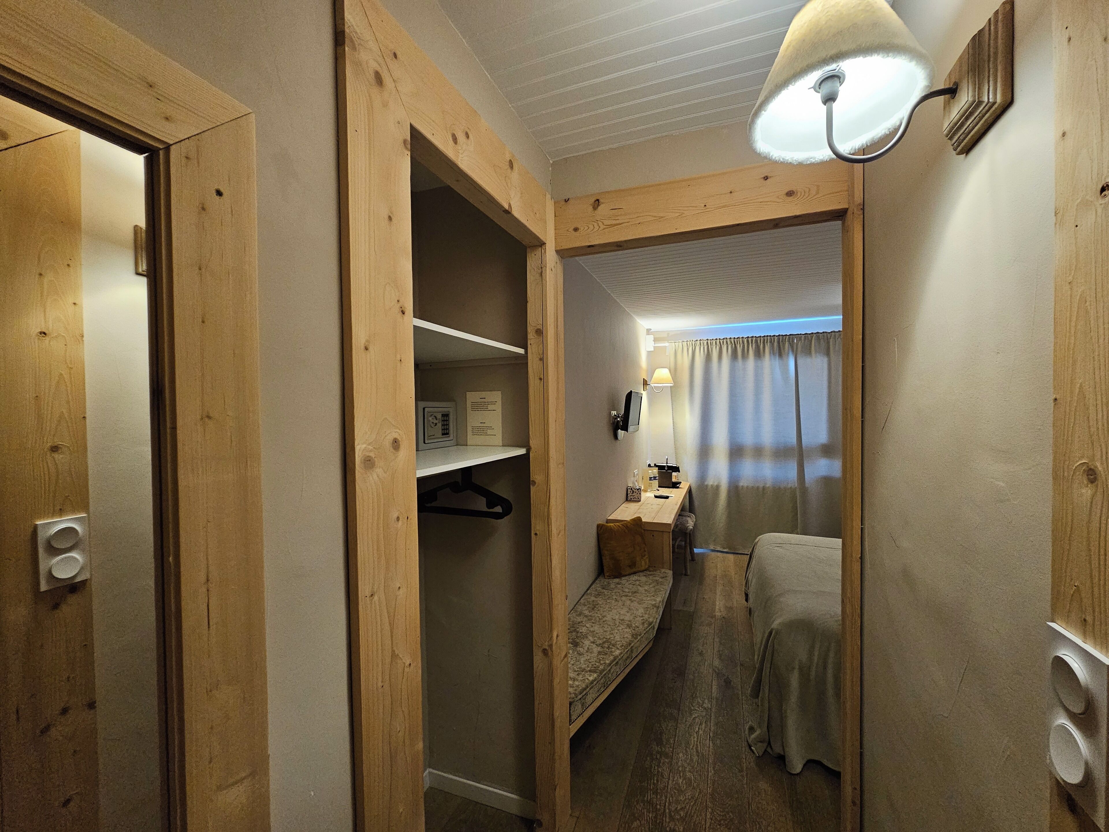 A narrow hallway leading to a bedroom with a bed, a desk, and a chair. There is a coat rack and a lamp on the wall.