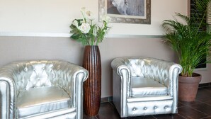 A reception area with two silver upholstered chairs, a tall vase with white flowers, a framed floral painting, and a potted plant. - Brit Hotel Confort Alexia La Souterraine (La Souterraine)