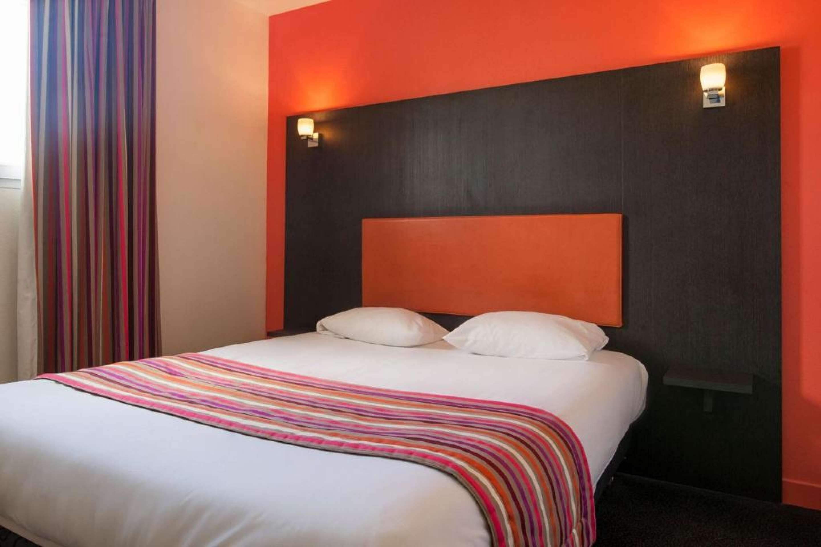 Een hotelkamer met een groot bed, twee kussens, een gestreepte deken en een donker hoofdeinde met twee wandlampen.