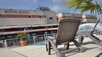 Un espace aménagé au bord de la piscine avec des chaises longues, un restaurant appelé « ALEXIA » en arrière-plan et une enseigne d’hôtel au-dessus.