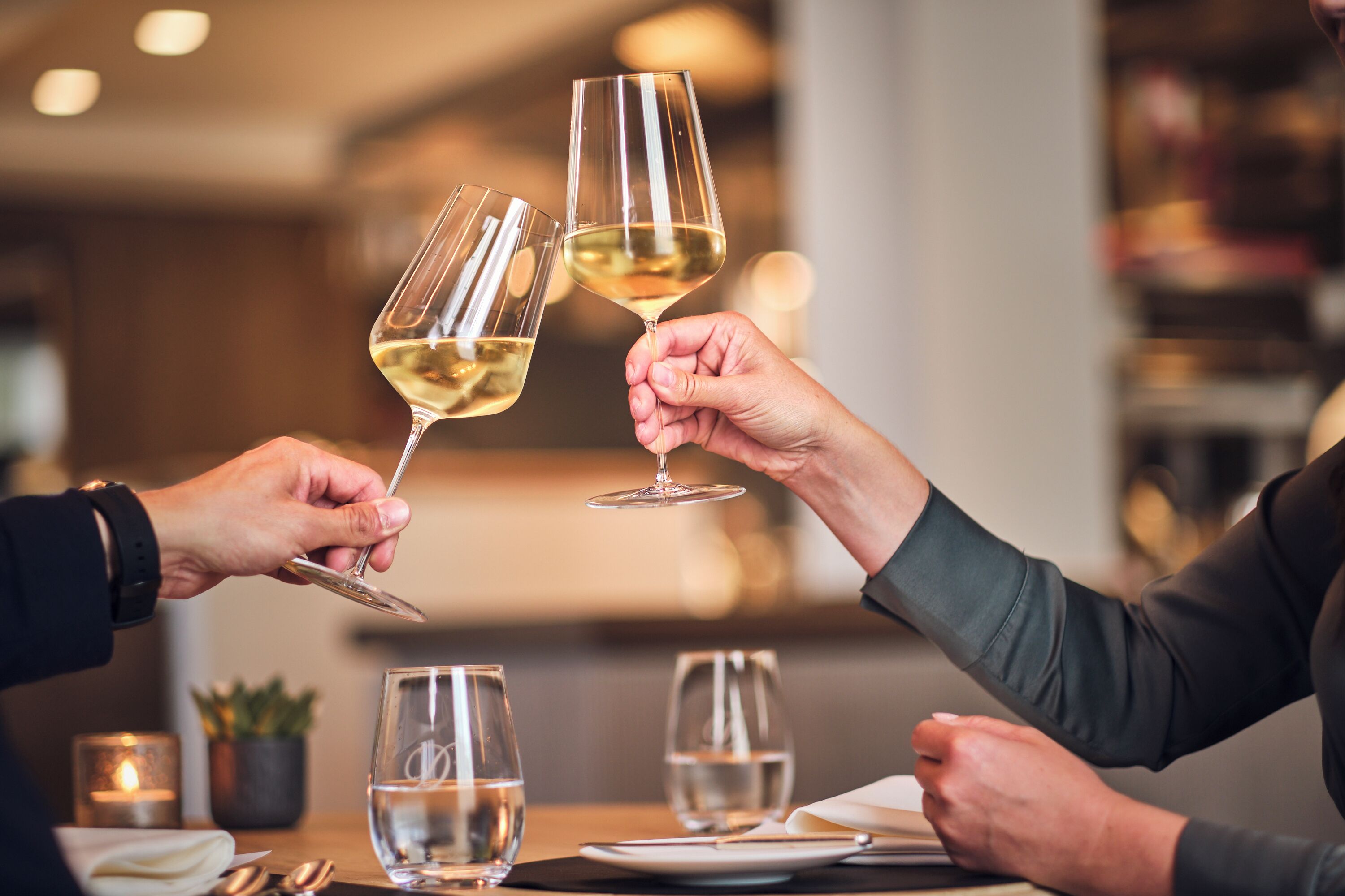 Zwei Personen stoßen mit Weingläsern an einem Tisch an, der mit Wassergläsern und einer Kerze bestückt ist, in einem schwach beleuchteten Restaurant.