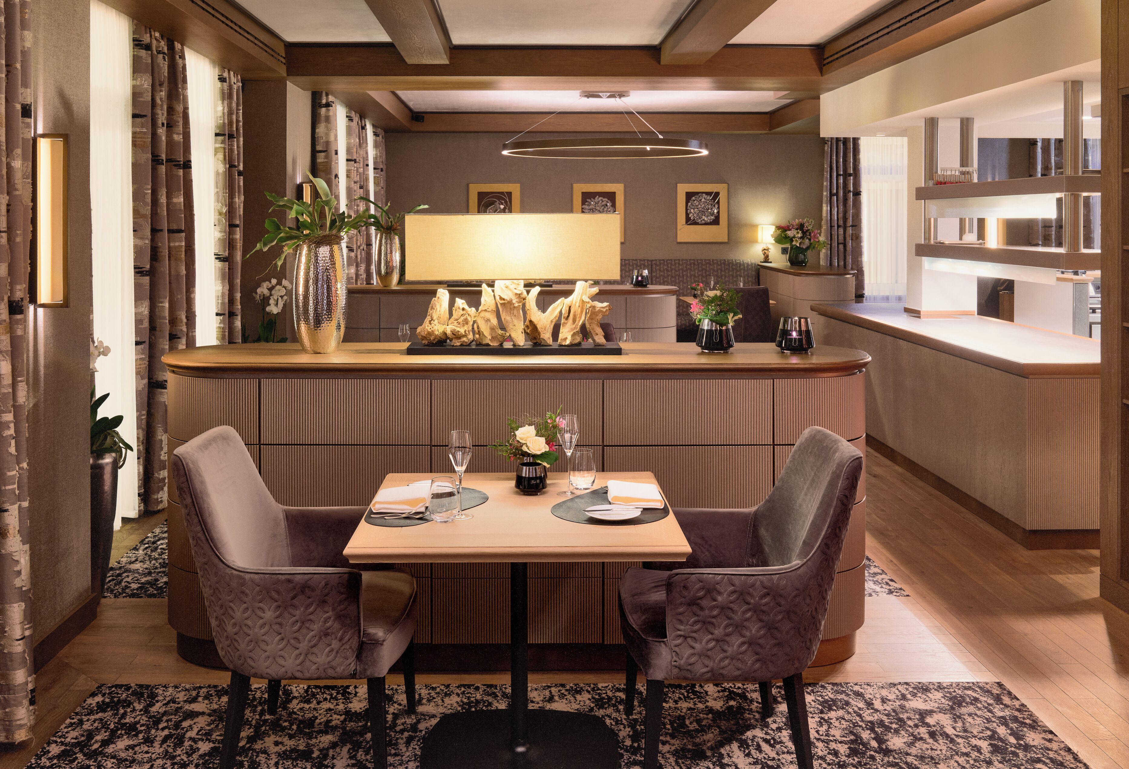 A modern hotel lobby with a reception desk, a dining area with two chairs and a table, and a kitchen area in the background.