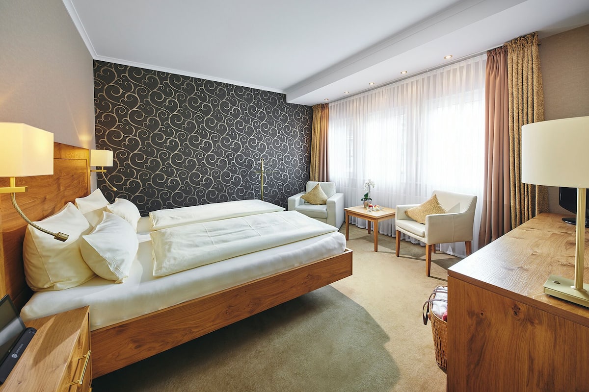 A hotel room with a large bed, two bedside lamps, a desk, a chair, and a patterned wall.