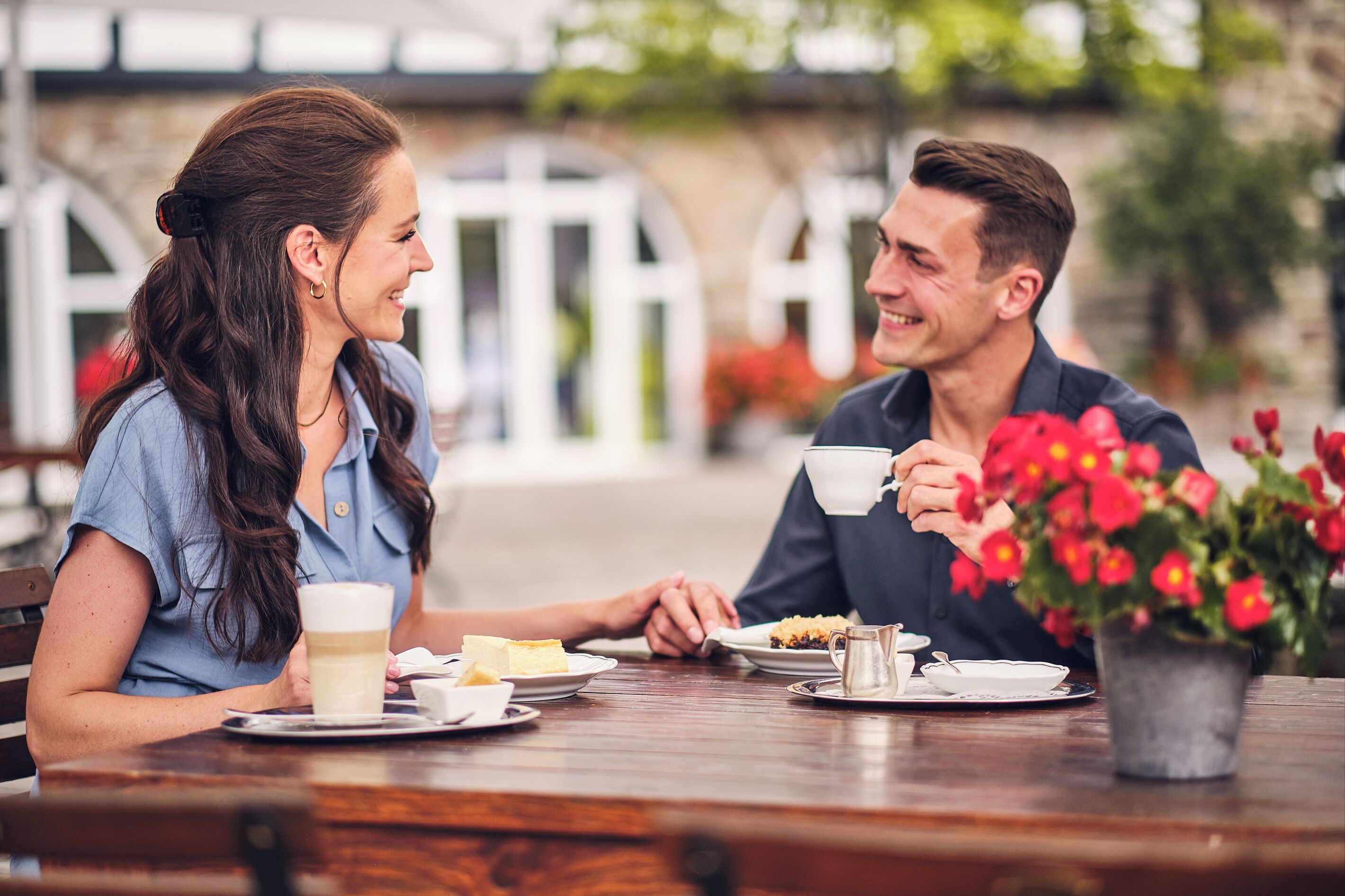 Two people sitting at a wooden table with coffee cups, plates of food, and a potted plant with red flowers.