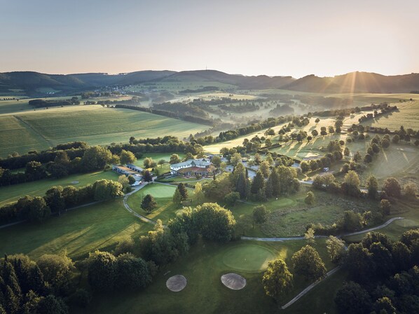 Une vue aérienne d’un terrain de golf avec un club-house, entouré de collines douces et de vastes champs.