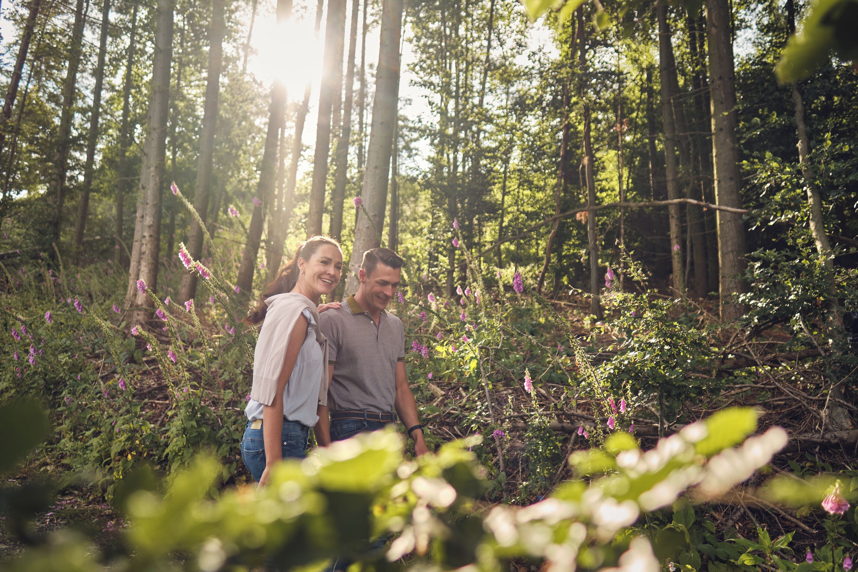 A couple standing in a forested area with sunlight filtering through the trees.