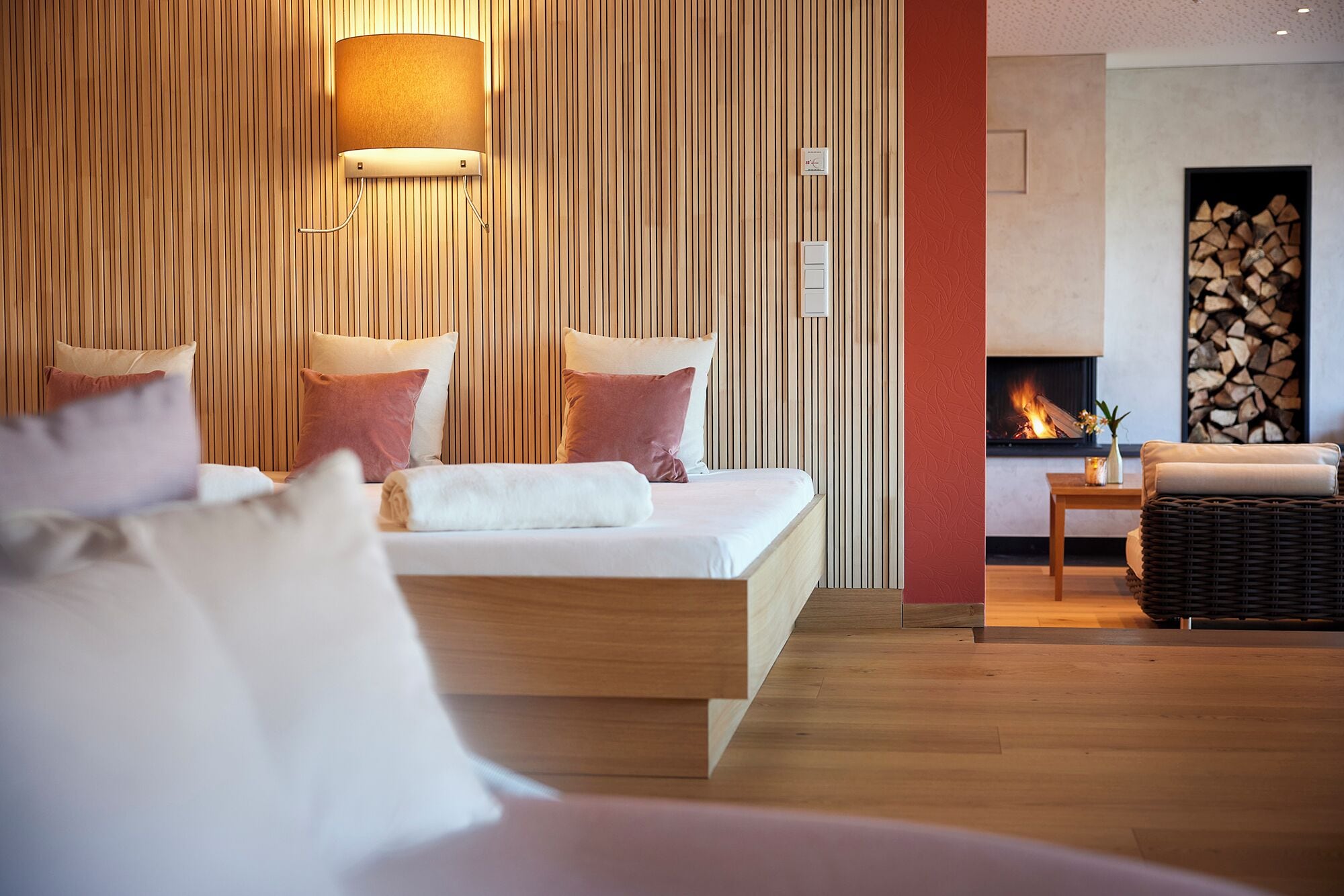 Een moderne hotelkamer met een wand van houtpanelen, een bed met witte beddengoed en roze kussens, een wandlamp en een open haard met brandhout.