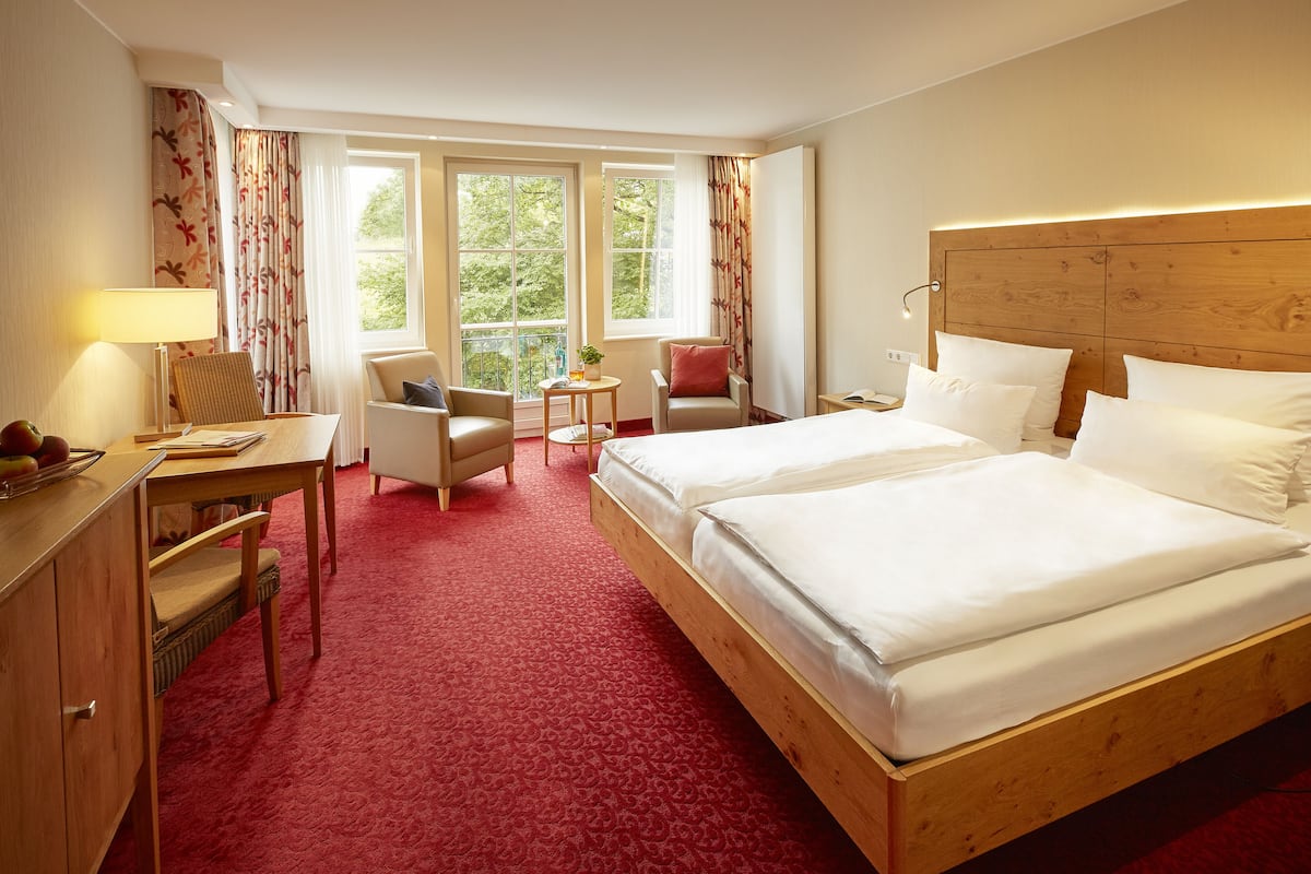 A hotel room with a large bed, a desk with a lamp, two chairs, a small table with a plant, and a window with curtains.