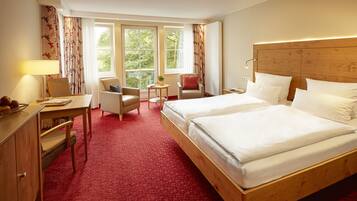 A hotel room with a large bed, a desk with a lamp, two chairs, a small table with a plant, and a window with curtains.