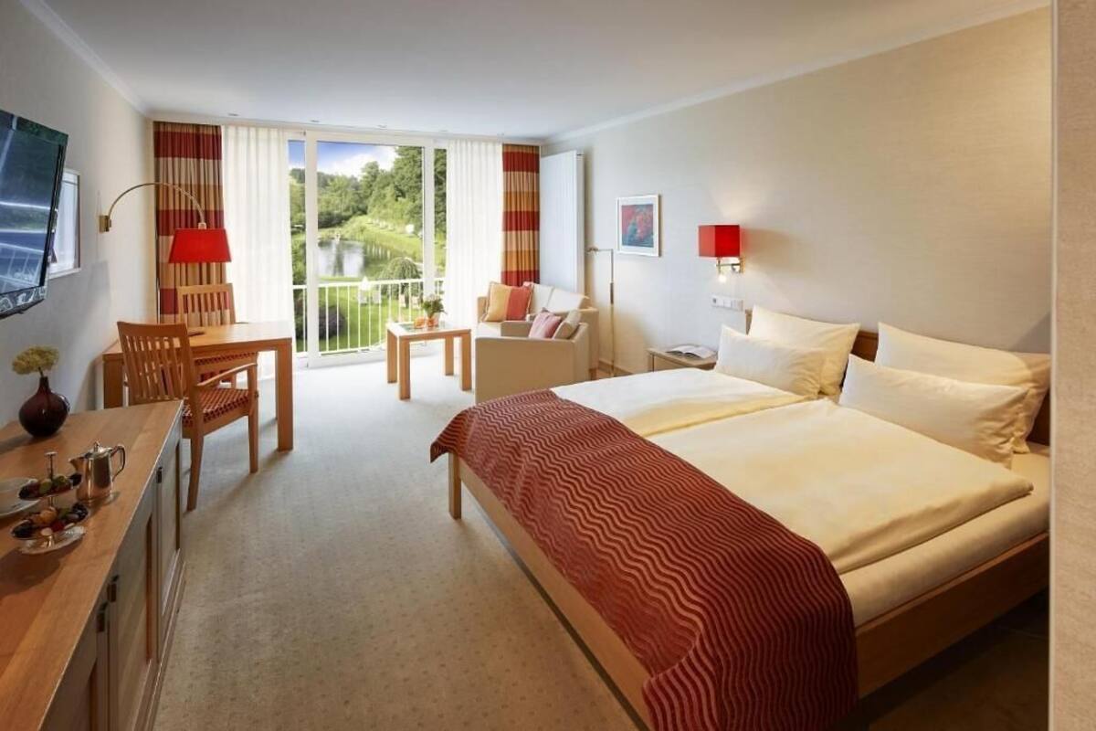 A hotel room with a bed, a desk, a chair, a small table, and a balcony with curtains.