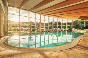 Una piscina interior con paredes de vidrio transparente, vigas de madera y suelo de baldosas.