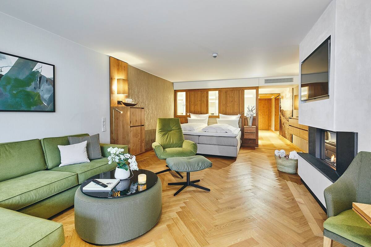 A spacious living area with a green sofa, a round coffee table, and a green armchair. A bed with white bedding is visible in the background.